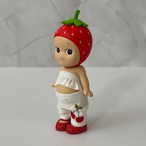 May include: A figurine of a smiling character wearing a red strawberry hat with green leaves, a white ruffled top, and white pants with cherry designs. The character has red shoes.