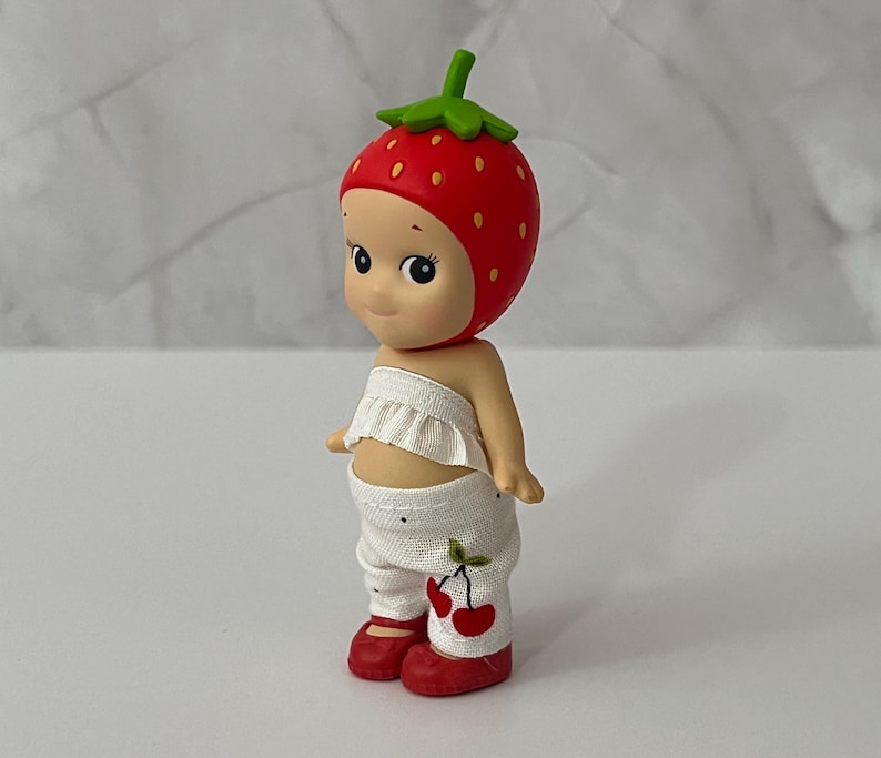 May include: A figurine of a smiling character wearing a red strawberry hat with green leaves, a white ruffled top, and white pants with cherry designs. The character has red shoes.