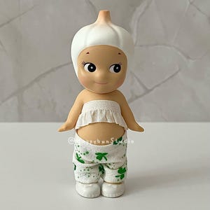 May include: A whimsical figurine of a baby-like character wearing a white garlic bulb hat and a matching ruffled top. The figurine is wearing white pants with green shamrock patterns and white shoes. The figurine has a smiling face and is a unique collectible.