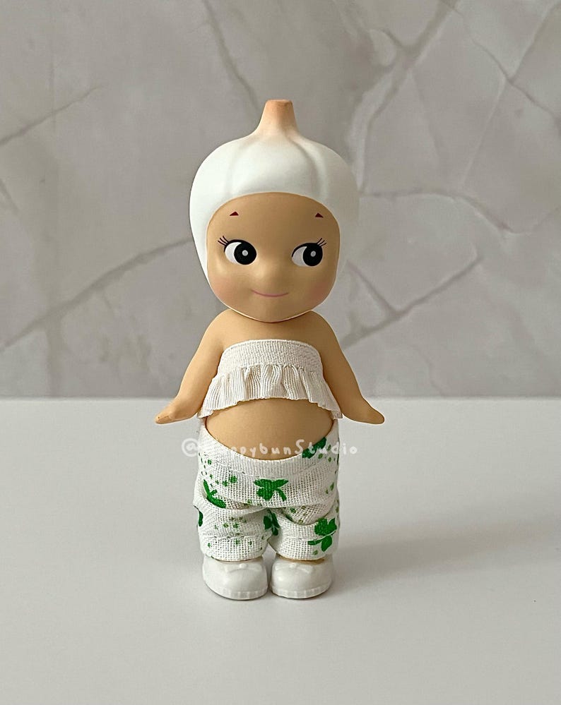 May include: A whimsical figurine of a baby-like character wearing a white garlic bulb hat and a matching ruffled top. The figurine is wearing white pants with green shamrock patterns and white shoes. The figurine has a smiling face and is a unique collectible.