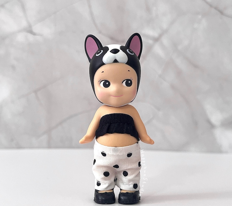 K&ouml;nnte beinhalten: Eine kleine Figur mit einer schwarz-wei&szlig;en Franz&ouml;sische Bulldogge-M&uuml;tze, einem schwarzen, ger&uuml;schten Schlauchoberteil und einer wei&szlig;en Hose mit schwarzen Punkten. Die Figur hat helle Haut, gro&szlig;e dunkle Augen und tr&auml;gt kleine schwarze Schuhe.