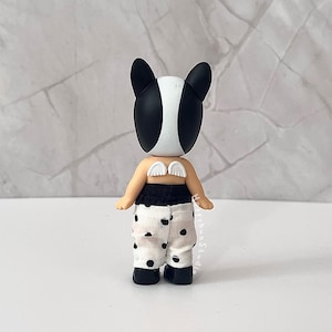 K&ouml;nnte beinhalten: Eine kleine Figur mit einem schwarz-wei&szlig;en tier&auml;hnlichen Kopf, kleinen wei&szlig;en Fl&uuml;geln auf dem R&uuml;cken und einer wei&szlig;en Hose mit schwarzem Punktemuster. Die Figur hat einen gebr&auml;unten Hautton und tr&auml;gt schwarze Schuhe und einen schwarzen Bund.
