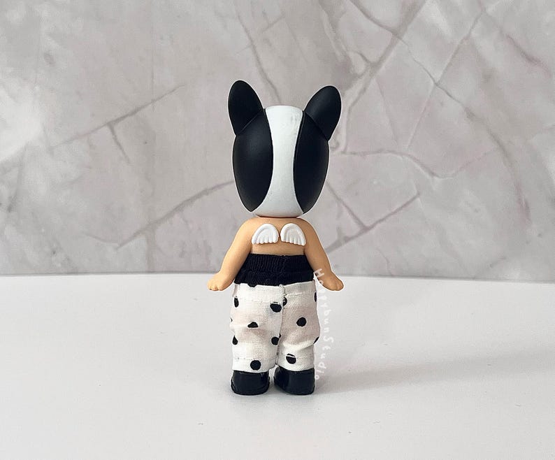 K&ouml;nnte beinhalten: Eine kleine Figur mit einem schwarz-wei&szlig;en tier&auml;hnlichen Kopf, kleinen wei&szlig;en Fl&uuml;geln auf dem R&uuml;cken und einer wei&szlig;en Hose mit schwarzem Punktemuster. Die Figur hat einen gebr&auml;unten Hautton und tr&auml;gt schwarze Schuhe und einen schwarzen Bund.