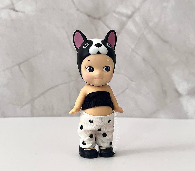 K&ouml;nnte beinhalten: Eine kleine Figur mit einer schwarz-wei&szlig;en Franz&ouml;sische Bulldogge-Kopfbedeckung, einem schwarzen tr&auml;gerlosen Oberteil und einer wei&szlig;en Hose mit schwarzen Punkten. Die Figur hat helle Haut, gro&szlig;e dunkle Augen und tr&auml;gt kleine schwarze Schuhe.