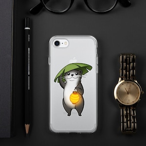 May include: A clear phone case with a cartoon otter wearing a green leaf hat and holding a yellow lantern. The otter is smiling and has a light gray body with dark gray accents.