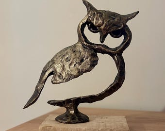 May include: A bronze-colored owl sculpture with a textured surface, perched on a wooden base. The owl has pointed ear tufts and a curved body, giving it a unique, artistic appearance. The sculpture is a decorative item.