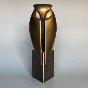 Può includere: Una scultura a forma di gufo color bronzo con un design geometrico. Il gufo ha un corpo aerodinamico e una base quadrata. La scultura è alta circa 30 cm e ha una finitura metallica, adatta all'arredamento della casa.