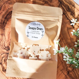 May include: A kraft paper bag with a clear window displaying flower-shaped lavender travel soaps. The bag has a circular label that reads "Splendidity Soapy Ones Lavender Travel Soap 15 Pieces". The soaps are white with brown specks. A sprig of white flowers is in the background.