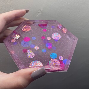 May include: A pink hexagonal coaster with a glittery surface and colorful iridescent circles.