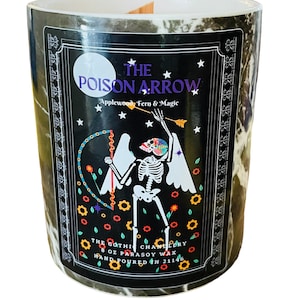 Puede incluir: Un tarro de vela de mármol blanco y negro con una etiqueta negra. La etiqueta presenta un esqueleto con alas sosteniendo una flecha, rodeado de estrellas y flores. El texto en la etiqueta dice "The Poison Arrow Applewood Fern & Magic The Gothic Chanllery 8 oz Parasoy Wax Hand Poured on 21146".