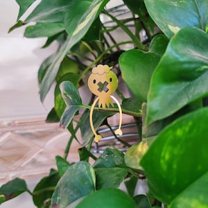 May include: A gold-coloured metal figurine of a person with a cross for a mouth, sitting on a green plant.