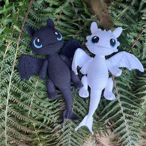 Op de afbeelding: Twee gehaakte draken knuffels, een zwarte en een witte, met blauwe ogen. De knuffels zijn geposeerd met hun vleugels uitgespreid en zitten op een bed van groene varens.