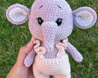 Può includere: Un giocattolo di peluche a forma di elefante lavorato a uncinetto in rosa e bianco. L'elefante indossa un vestito lavorato a uncinetto rosa e bianco con un bordo a balze. L'elefante ha grandi occhi neri e un piccolo naso rosa lavorato a uncinetto.
