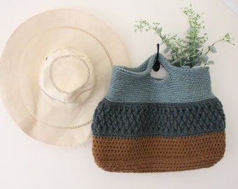 May include: A crocheted tote bag with a blue, green, and brown color scheme. The bag has a single handle and is hanging on a hook. A straw hat is hanging on the wall behind the bag.