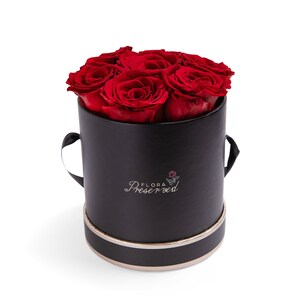 May include: A black box with a gold trim, containing a bouquet of preserved red roses. The box has two handles and the text "FLORA Preserved" on the front.