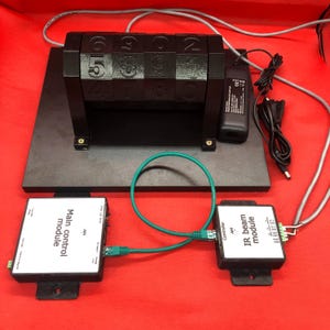 Puede incluir: Una pantalla digital negra con bloques numerados, conectada a dos módulos blancos etiquetados como "Main control module" e "IR beam module". Los módulos están conectados por un cable verde. La pantalla está sobre una base negra sobre un fondo rojo.