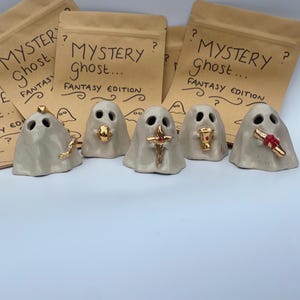 May include: Five small, light grey ceramic ghost figurines, each holding a different gold-coloured accessory, including a crown, orb, sword, chalice, and scroll. Behind them are brown paper pouches labelled MYSTERY ghost... FANTASY EDITION.