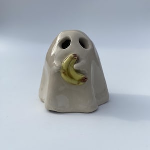 May include: A small, glossy, light beige ceramic ghost figurine with two dark, hollow eye sockets. The ghost is holding a small, curved, yellow banana with brown spots in its centre, featuring a smooth, reflective glaze finish throughout.