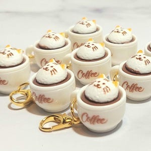 May include: A collection of miniature white coffee cups with gold-colored handles and keychains. Each cup is filled with brown liquid and topped with a white cat-shaped design. The word "Coffee" is written on the side of each cup.
