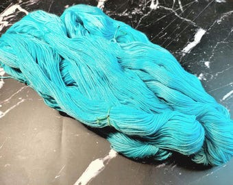 May include: A skein of vibrant turquoise yarn, tightly wound and secured with a green band. The yarn is a bright, solid color, perfect for knitting or crocheting projects. The yarn is displayed on a dark surface.