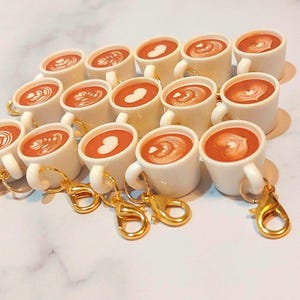 May include: A collection of miniature white ceramic coffee mugs with gold-colored keychains. Each mug is filled with a brown liquid and topped with latte art. The latte art designs include hearts and swirls. The mugs are arranged on a white marble surface.