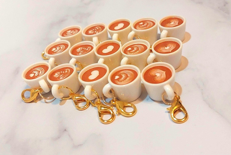 May include: A collection of miniature white ceramic coffee mugs with gold-colored keychains. Each mug is filled with a brown liquid and topped with latte art. The latte art designs include hearts and swirls. The mugs are arranged on a white marble surface.