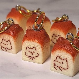 May include: A collection of square-shaped bread loaf charms, each featuring a golden metal lobster clasp. The charms have a light cream-colored base with a textured, browned top crust and a small, simple line-art drawing of a cat face on the front.