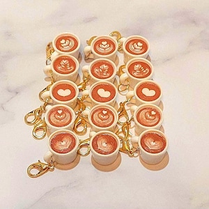 May include: A collection of miniature coffee cup keychains. Each white cup features a latte art design in shades of brown and tan. The keychains have gold-colored clasps.