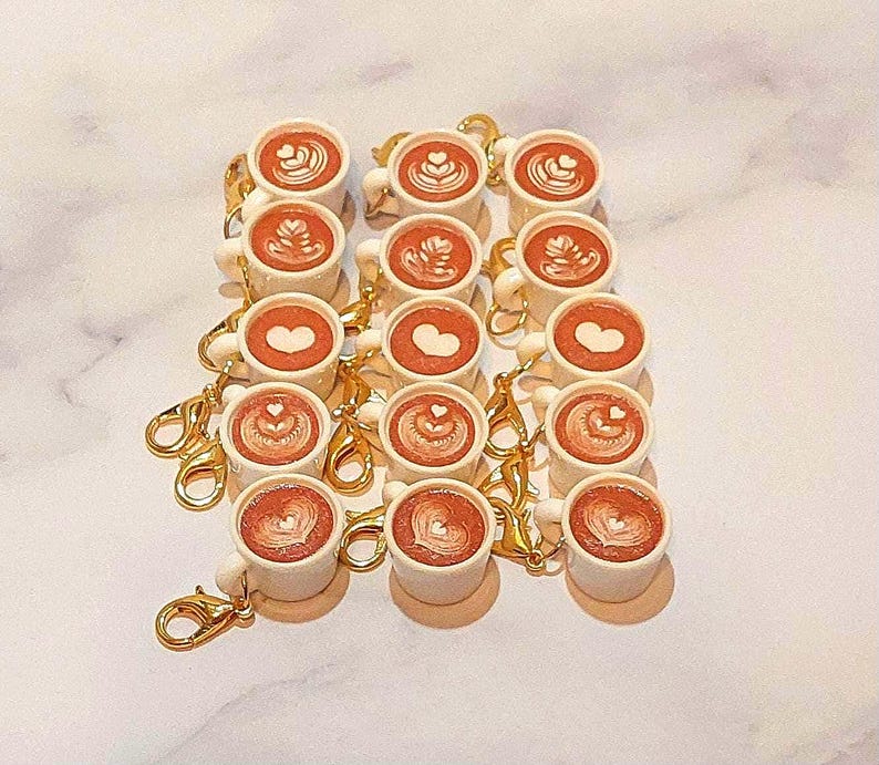 May include: A collection of miniature coffee cup keychains. Each white cup features a latte art design in shades of brown and tan. The keychains have gold-colored clasps.