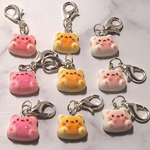 May include: A collection of eight small charms featuring stylized cat faces in shades of pink, orange, and white. Each charm is attached to a silver-toned metal lobster clasp. The cat faces have simple facial features and small paws.