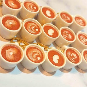 May include: A collection of miniature white ceramic coffee cups, each filled with a reddish-brown liquid and topped with latte art. The latte art designs include hearts and swirls. The cups are connected by a gold chain.