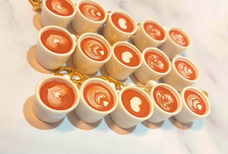May include: A collection of miniature white ceramic coffee cups, each filled with a reddish-brown liquid and topped with latte art. The latte art designs include hearts and swirls. The cups are connected by a gold chain.