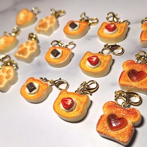 May include: A collection of small, golden-brown toast-shaped charms with gold-tone lobster clasps. The charms feature various toppings including red heart-shaped centers, sliced strawberries with white cream, and dark chocolate squares or paw prints.