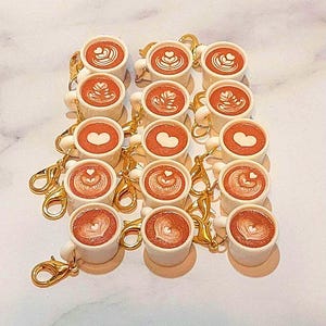May include: A collection of miniature white coffee cup keychains with gold-colored clasps. Each cup is filled with a brown liquid and topped with latte art in the shape of hearts and other designs. The keychains are arranged in neat rows.