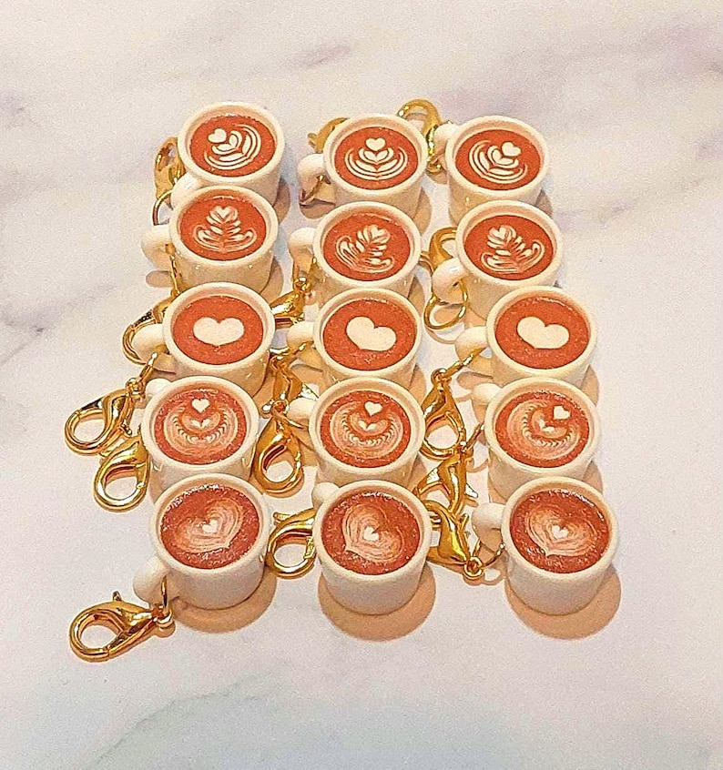 May include: A collection of miniature white coffee cup keychains with gold-colored clasps. Each cup is filled with a brown liquid and topped with latte art in the shape of hearts and other designs. The keychains are arranged in neat rows.