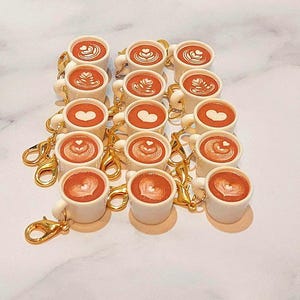 May include: A collection of miniature white coffee mugs with gold-colored clasps. Each mug features a brown coffee design with latte art, including hearts and swirls. The mugs are arranged in rows on a white surface.