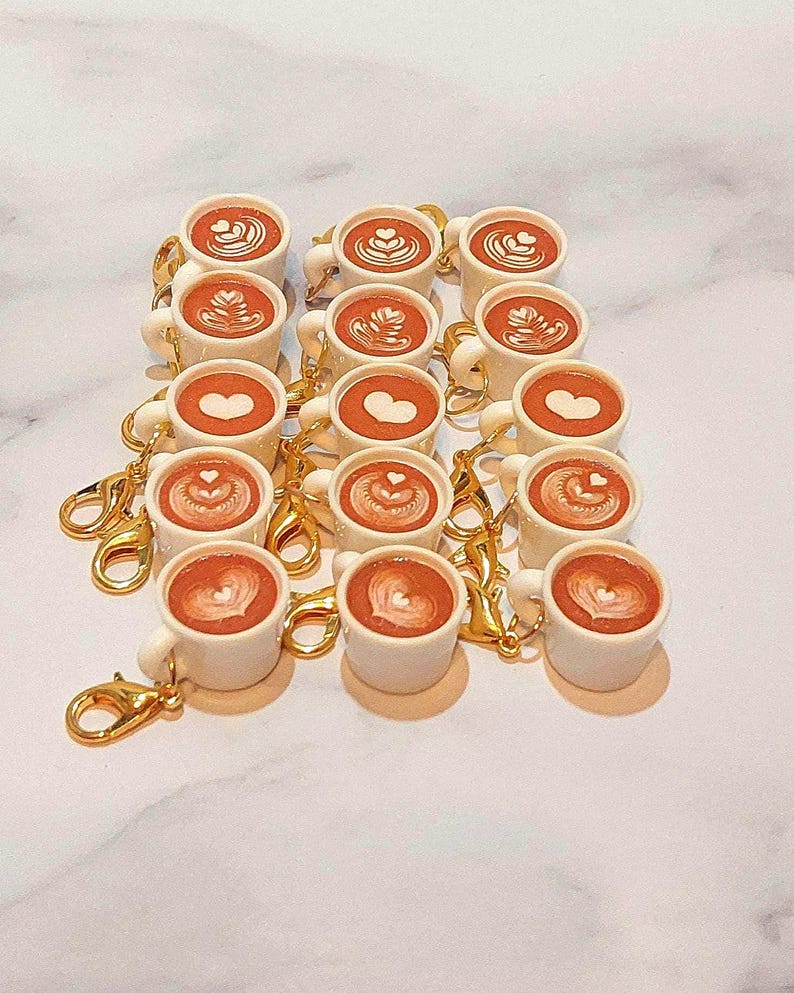 May include: A collection of miniature white coffee mugs with gold-colored clasps. Each mug features a brown coffee design with latte art, including hearts and swirls. The mugs are arranged in rows on a white surface.