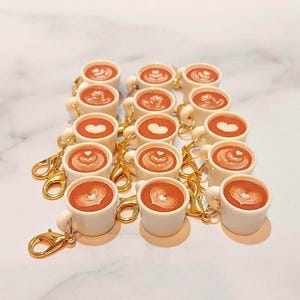 May include: A collection of miniature white coffee mugs with gold-colored clasps. Each mug is filled with a brown liquid and topped with latte art. The latte art designs include hearts and other patterns.
