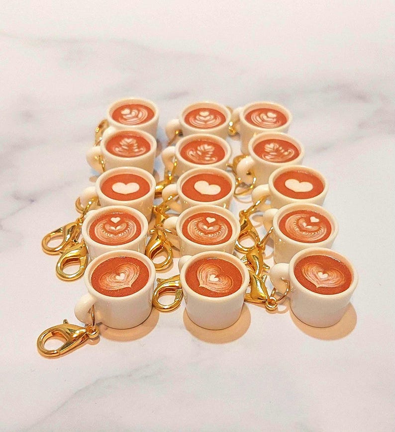 May include: A collection of miniature white coffee mugs with gold-colored clasps. Each mug is filled with a brown liquid and topped with latte art. The latte art designs include hearts and other patterns.