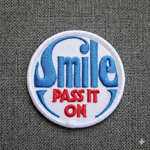 May include: Circular embroidered patch featuring the words "Smile" in blue and "Pass It On" in red. The patch has a white background and a white border. The design is circular.