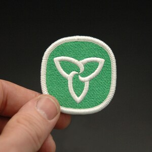 May include: A green and white embroidered patch featuring the Ontario provincial symbol, a stylized trillium flower. The patch has a white border and is held against a dark background.