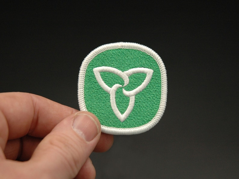 May include: A green and white embroidered patch featuring the Ontario provincial symbol, a stylized trillium flower. The patch has a white border and is held against a dark background.
