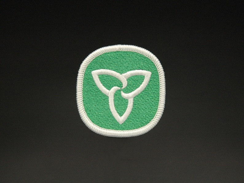 May include: A green and white embroidered patch featuring the Ontario provincial symbol, a stylized trillium flower. The patch has a white border and a green background. The design is a three-petal floral emblem.