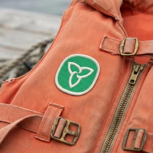 May include: Close-up of an orange life vest with a green and white emblem. The vest has a zipper, straps, and metal buckles. The background includes a rope and water.