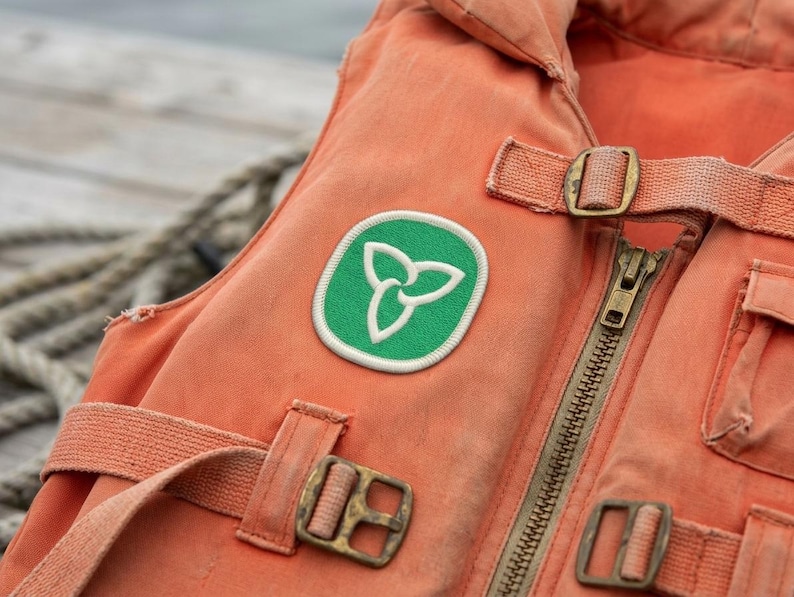 May include: Close-up of an orange life vest with a green and white emblem. The vest has a zipper, straps, and metal buckles. The background includes a rope and water.