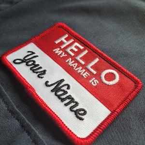 May include: Red and white fabric name tag with the text "HELLO MY NAME IS" and "Your Name" in black embroidery.
