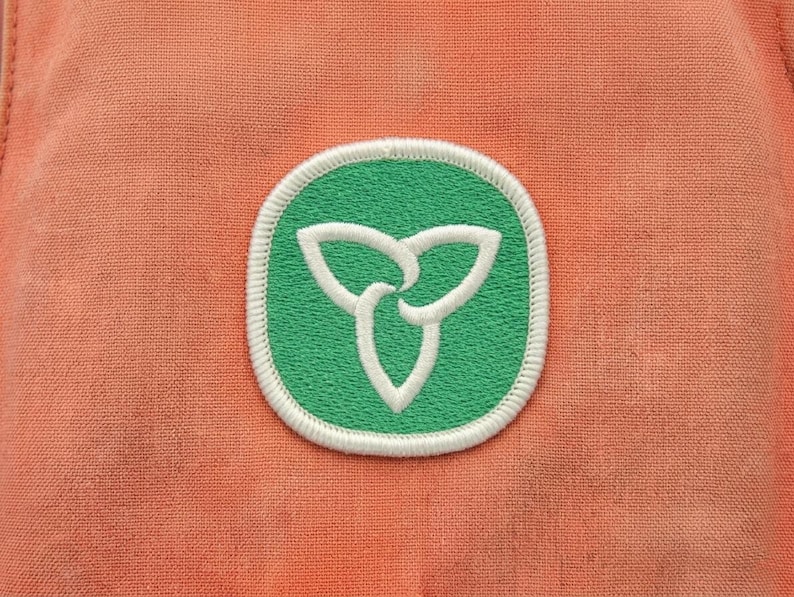 May include: Close-up of a patch featuring a green circle with a white, stylized three-leaf clover design. The patch is bordered with white stitching and set against a textured, peach-colored fabric.