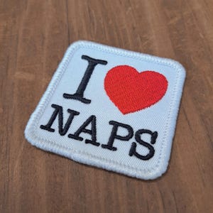May include: Square white patch with the words "I LOVE NAPS" in black, with a red heart replacing the word "love". The patch has a white border and is on a wooden surface.