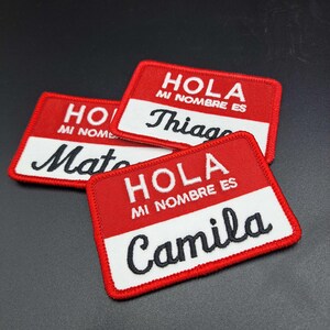 May include: Three red and white embroidered name patches. Each patch has the text "HOLA MI NOMBRE ES" in white, with a name in black script below. The names are "Mate", "Thiago", and "Camila".