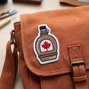 May include: An embroidered patch featuring a brown maple syrup bottle with a red maple leaf emblem. The patch is affixed to a rust-colored canvas messenger bag with a buckle closure and adjustable strap.